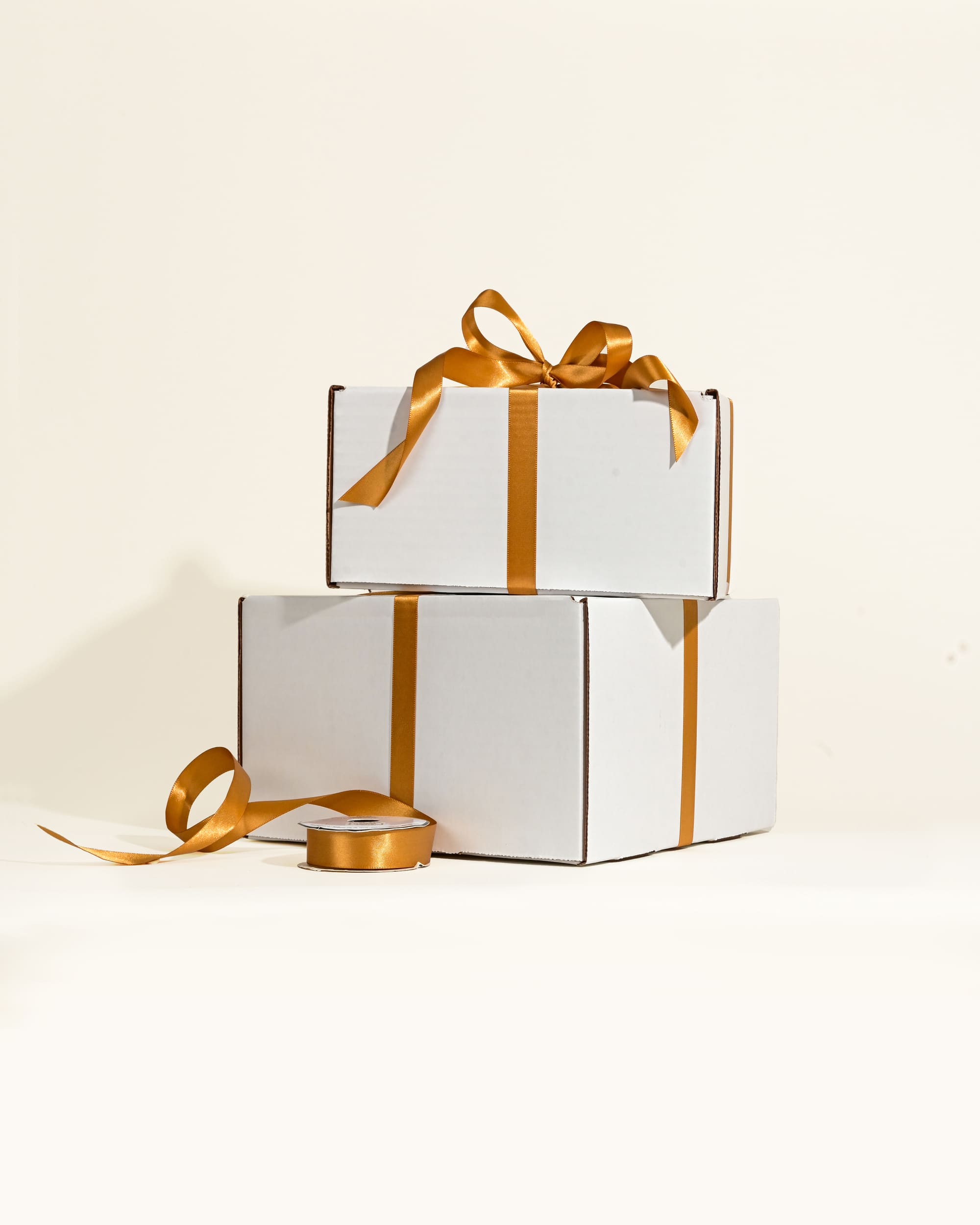 Two white gift boxes with gold ribbons stacked on a light beige background