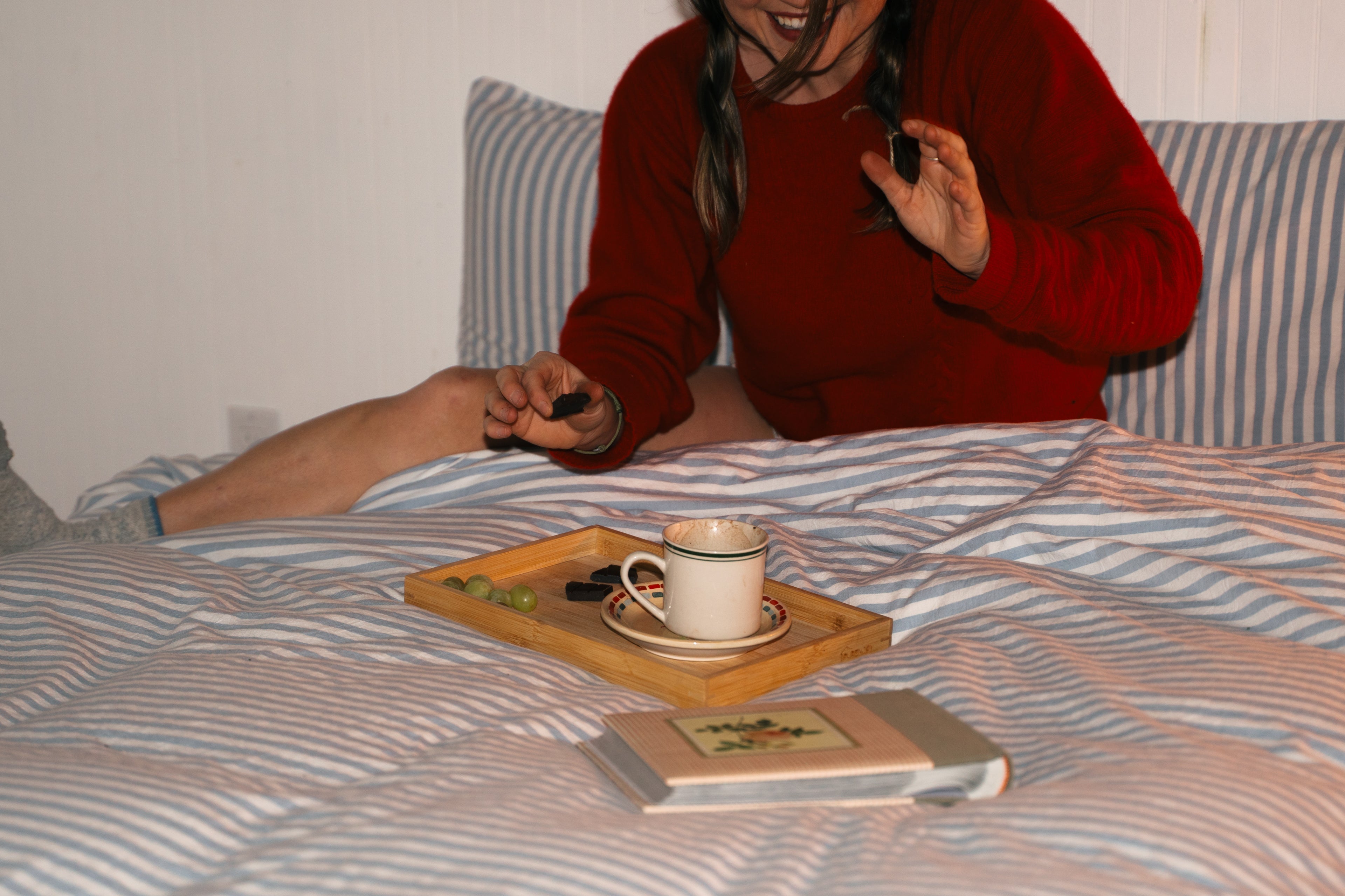 Person sitting on a bed with a tray holding a cup of hot cocoa and Seahorse craft chocolate, wearing a red sweater.